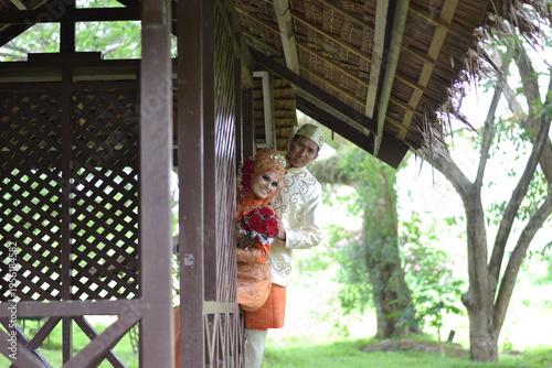 A happy indonesian couple in traditional orange and cream wedding attire posing playfully by a rustic wooden gazebo in a lush green garden