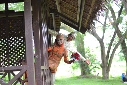 A happy indonesian couple in traditional orange and cream wedding attire posing playfully by a rustic wooden gazebo in a lush green garden
