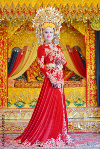 A portrait of a Minangkabau bride in a detailed red and gold traditional dress and elaborate headdress, posed against an ornate wedding backdrop