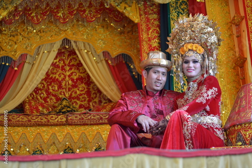 A happy Indonesian couple in traditional Minangkabau red and gold wedding attire sitting together on a decorative bridal throne