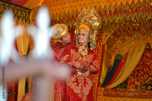 A smiling Indonesian couple in traditional red and gold attire on a lushly decorated wedding stage