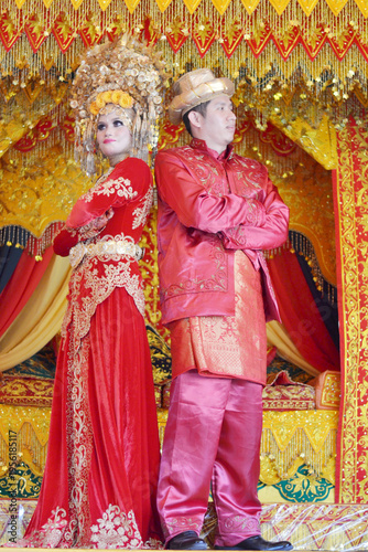 A stunning Acehnese couple in traditional red and gold wedding attire, posing elegantly against a vibrant, ornate ceremonial backdrop