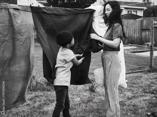 A mother and her child engage in a bonding activity by hanging laundry outdoors. The black and white image captures a moment of connection and everyday life.