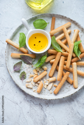 Grey plate with breadsticks, salt, olive oil and basil, vertical shot on a white stone background, middle close-up, high angle view