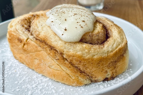 Fresh homemade cinnamon roll with creamy icing on a white plate, soft fluffy texture, cozy bakery style dessert.