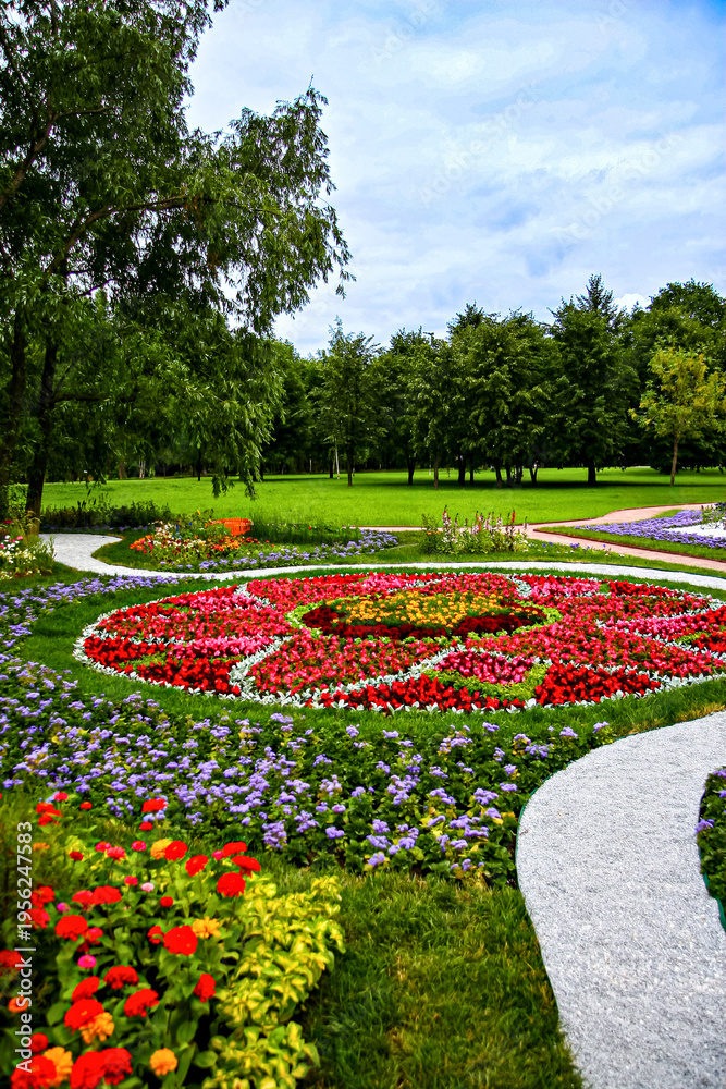 Naklejka premium Symmetrical circular flower arrangement surrounded by winding paths and greenery, showcasing a harmonious park design under a bright sky.
