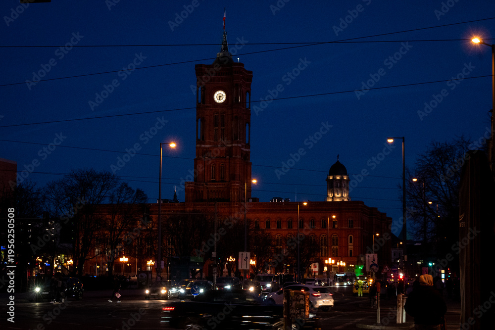 Fototapeta premium Nighttime view of a historic clock tower and surrounding architecture illuminated by warm lights against a deep blue sky.