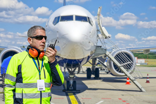 Airport ground crew worker coordinating operations with walkie-talkie during aircraft ground handling on ramp.