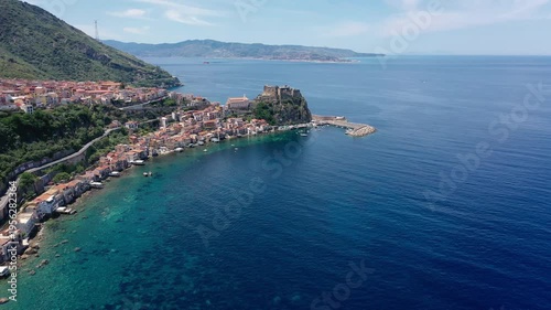 Slow aerial pan left along the coast of Scilla in Calabria showing a fortress on a promontory blue water rocks road and clear sky.