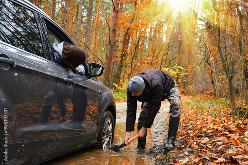 Fotografi Man digging mud to free stuck car on forest road while another person watches fr