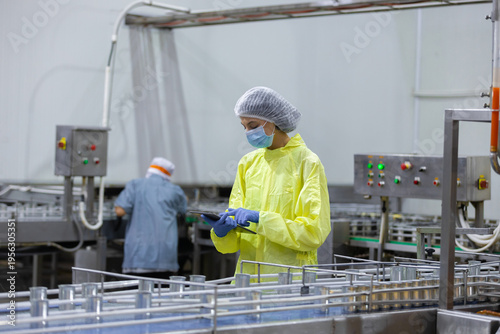 Quality control inspectors in protective clothing checking food production line in factory using clipboard to monitor hygiene standards, safety compliance and industrial processing efficiency.
