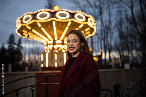 A woman in an amusement park with a carousel in the background is happy and smiling in the evening