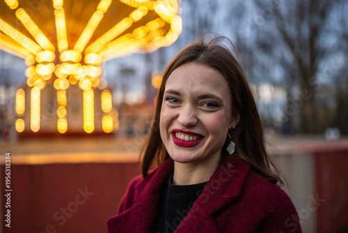 A woman in an amusement park with a carousel in the background is happy and smiling in the evening
