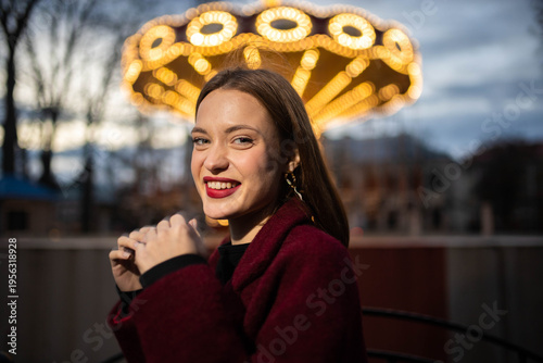 A woman in an amusement park with a carousel in the background is happy and smiling in the evening