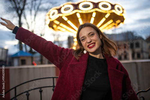 A woman in an amusement park with a carousel in the background is happy and smiling in the evening