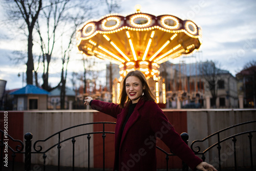 A woman in an amusement park with a carousel in the background is happy and smiling in the evening