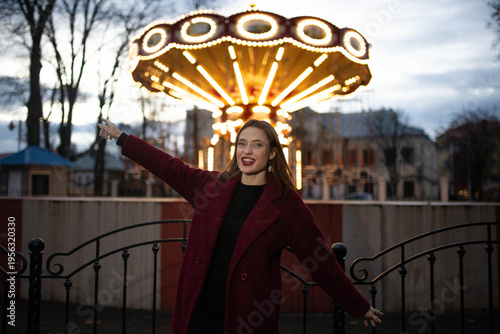 A woman in an amusement park with a carousel in the background is happy and smiling in the evening