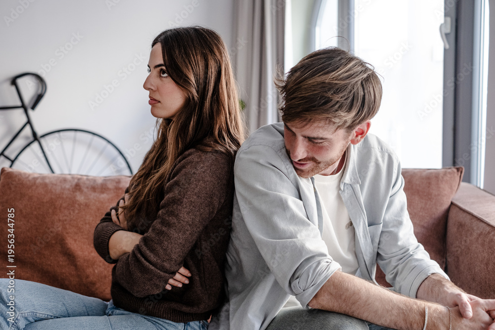 Obraz premium Young couple sitting back to back on couch having a dispute, expressing relationship conflict, unresolved problem or question, emotional distance, or communication problems in a modern living room.