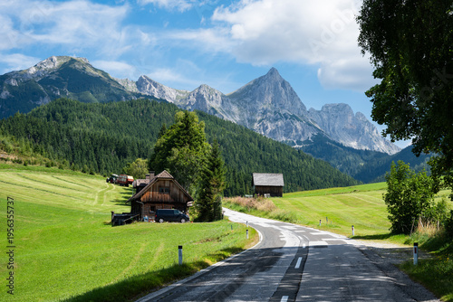 Photos A winding road leads past traditional wooden houses and green meadows toward the sharp, towering peaks of the Austrian Alps under a blue sky in Austria