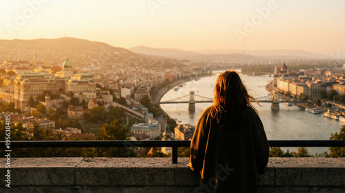 A person stands at a stone railing overlooking a city with a river and bridges at sunset, with hills and historic buildings in the background.