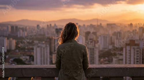 A person with long hair and a jacket stands on a balcony, looking out over a sprawling cityscape during a warm, golden sunset.