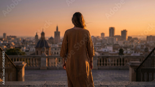 A woman in a brown dress stands with her back to the camera, gazing at a city skyline during sunset from a stone terrace with railings.