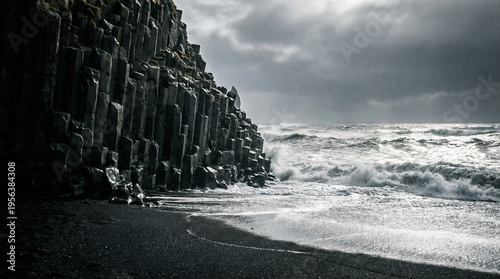 Photography Dark basalt rock formations rise sharply beside a turbulent ocean under a cloudy sky with waves crashing on a black sand beach