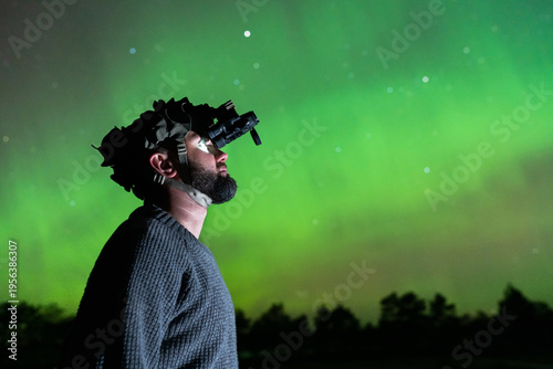 Side profile of a man using a professional night vision monocular device under a glowing green Aurora Borealis sky.