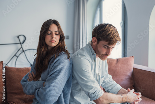фотографія A young couple sits back to back on a sofa, both looking upset and withdrawn after a disagreement; the image conveys tension, distance, emotional conflict and relationship strain at home
