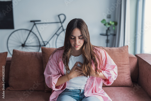 Young woman sits on a sofa clutching her chest and wincing, conveying chest pain, anxiety and possible heart-related discomfort in a casual home setting for health, medical and wellness topics.