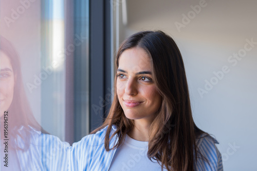 Young woman stands by a window smiling softly as she gazes outside, her reflection visible in the glass; natural light highlights a calm, hopeful and thoughtful mood.