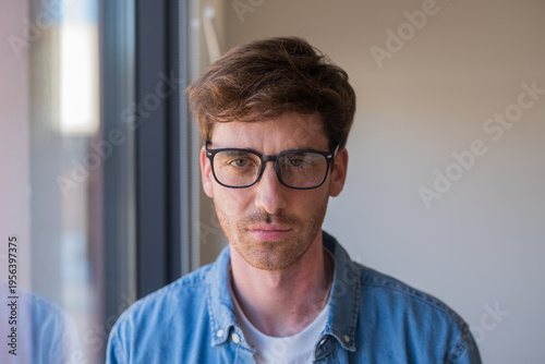 Thoughtful young man wearing glasses and a denim shirt looks toward the camera by a window, conveying introspection, focus and quiet confidence in a natural indoor portrait.