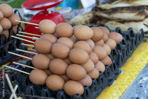 Close-up of numerous grilled brown eggs neatly arranged on wooden skewers and displayed in egg trays at a vibrant Thai street food market.