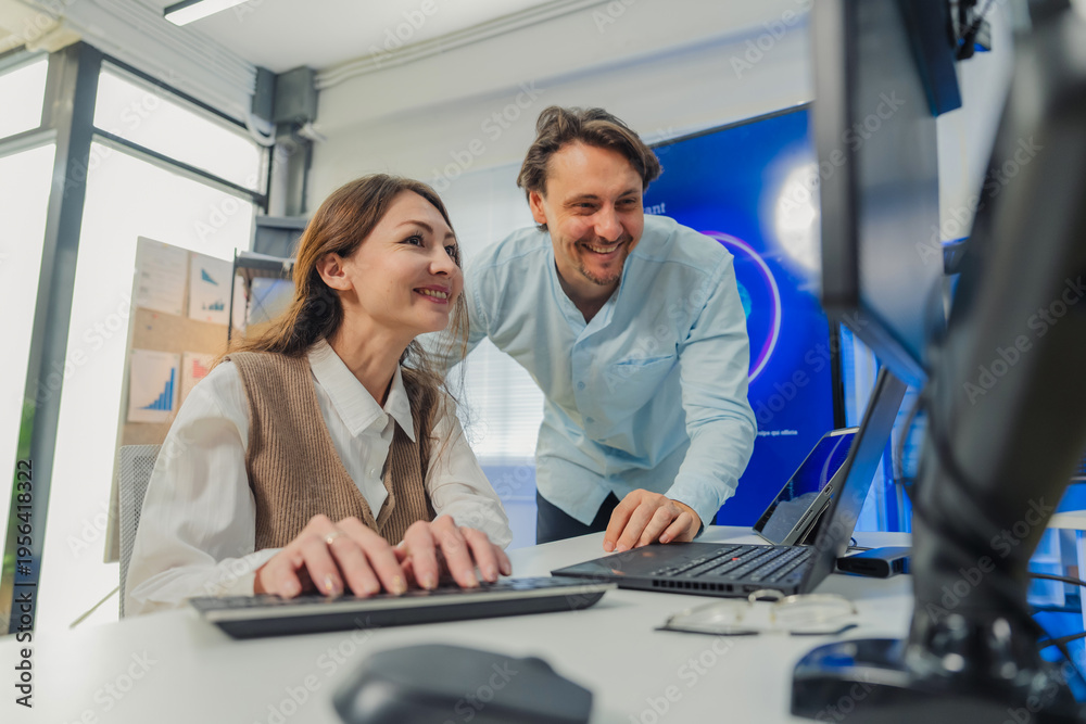 Fototapeta premium female developer in vest codes on laptop while male colleague in blue shirt leans over pointing at AI data visualizations on multiple monitors in modern tech office with large AI Assistant screen