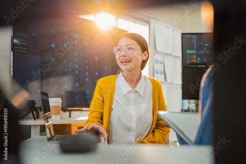 collaborative female AI developer in yellow sweater and glasses discusses code and neural network diagrams on large monitor while taking notes on tablet in modern tech office, with team members listen
