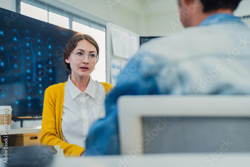 collaborative female AI developer in yellow sweater and glasses discusses code and neural network diagrams on large monitor while taking notes on tablet in modern tech office, with team members listen