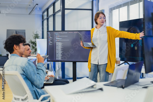 confident female AI team leader in yellow cardigan and glasses presents neural network architecture and code on large monitor while explaining to seated developers with laptops and tablets in office
