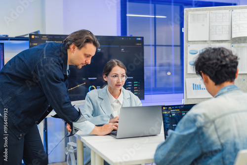 collaborative AI development team in modern tech office: young male developer coding on laptop, female team leader in light blue blazer pointing at code and neural network visualization 