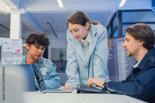 collaborative AI development team in modern tech office: young male developer coding on laptop, female team leader in light blue blazer pointing at code and neural network visualization 