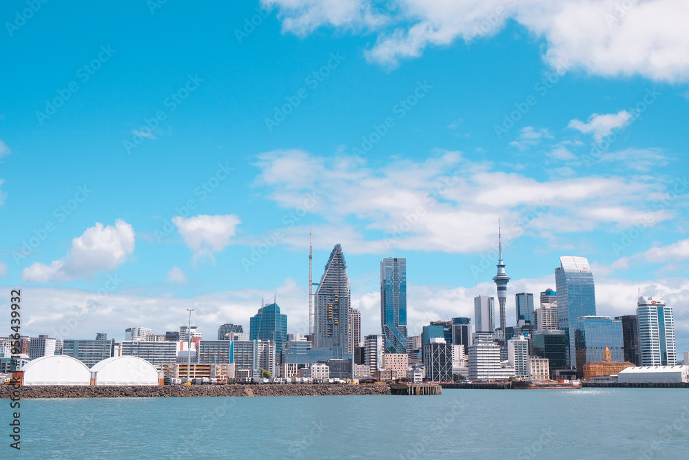 Fototapeta premium Auckland, New Zealand: The city skyline and harbor edge seen from a distance on the water with a wide blue sky overhead