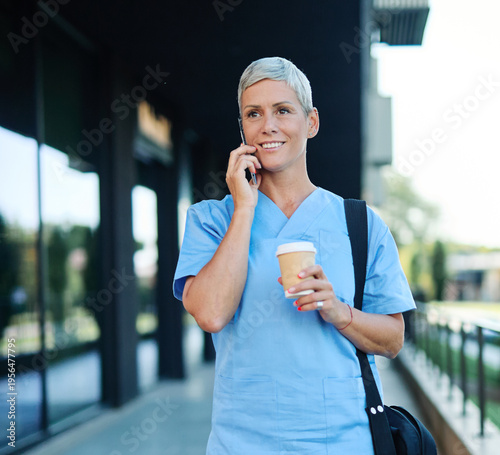 Nurse in scrubs enjoying coffee and using a mobile phone outside a modern medical facility during a sunny day