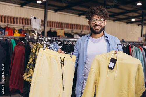 Young smiling Arab man holding a yellow hoodie and sweatpants set, shopping for fashion in a modern clothing store
