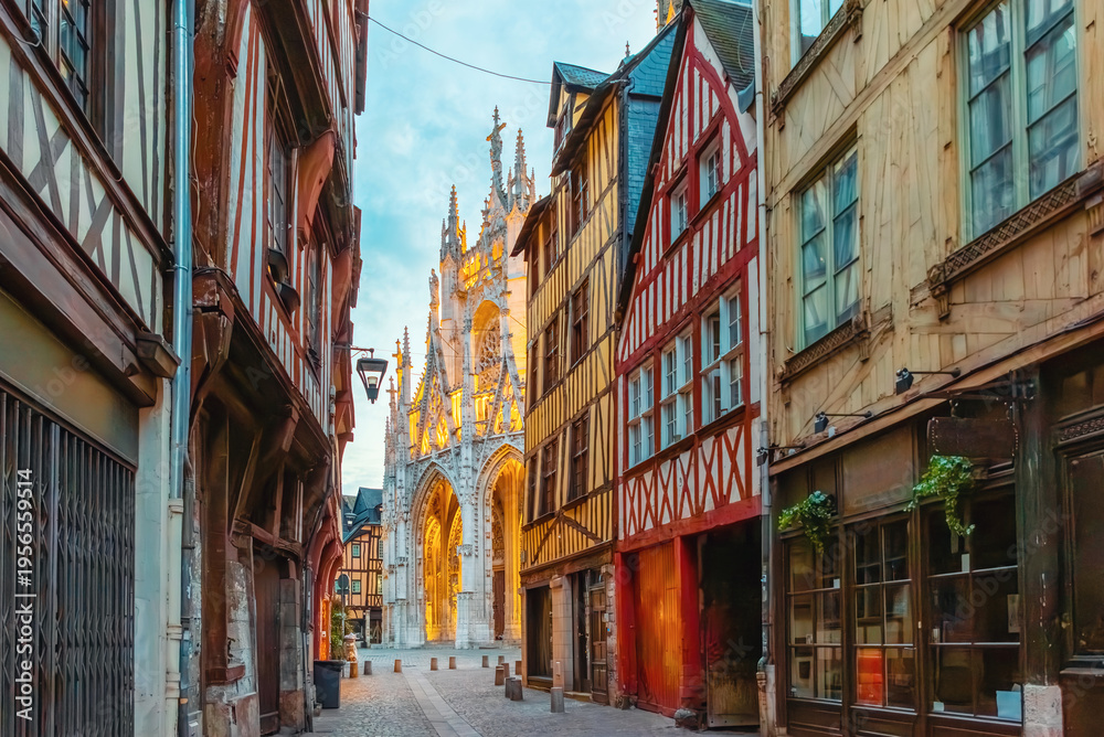 Fototapeta premium Narrow medieval street in Rouen with half timbered houses leading to illuminated Gothic cathedral facade at sunrise, Normandy, France. Historic architecture and European travel destination