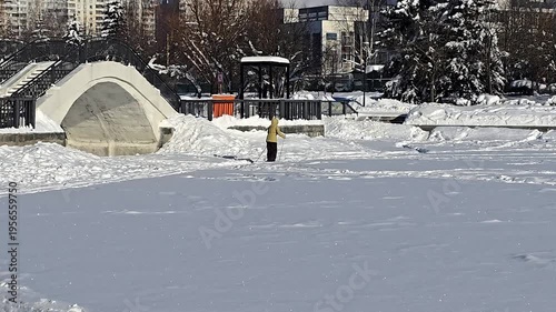 Middle-aged woman with fair skin in a yellow jacket cross-country skis on a snowy field near an arch bridge in a winter city park.