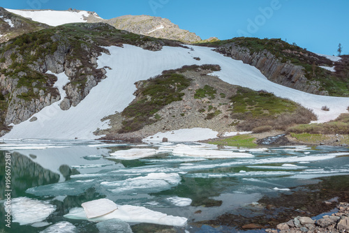 Pieces of ice float in mountain lake among sharp rocks, sheer crags and snowf...