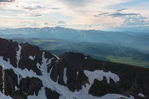 Aerial view above black sharp rocky ridge to sunlit green meadows and dark si...