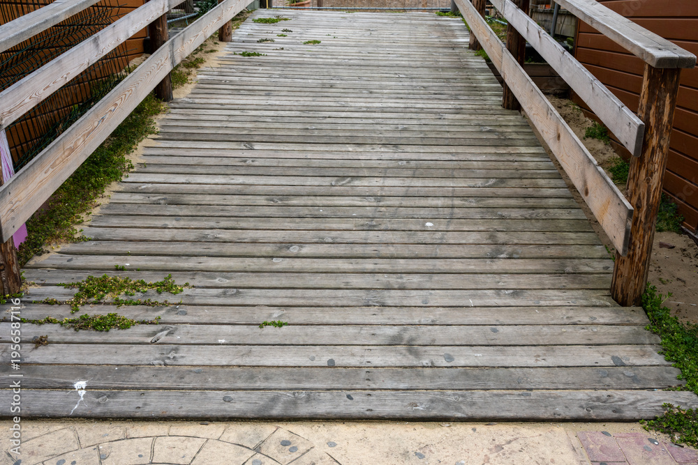 Fototapeta premium a weathered grey wooden boardwalk ramp with railings, featuring natural wood textures and small plants growing through the slats, captured under bright daylight.
