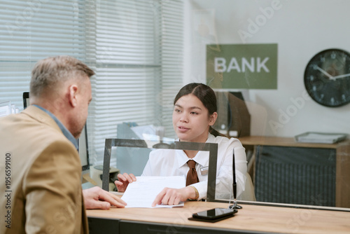 Schilderij op canvas Caucasian man speaking with young adult Asian bank teller at branch counter, wom