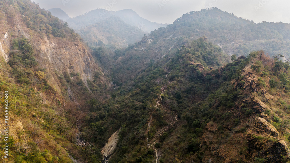 Fototapeta premium Aerial view of the Song River in Maldevta near Dehradun, Uttarakhand, showing low water flow in winter with a village house, greenery, and dirt roads across the mountain landscape.