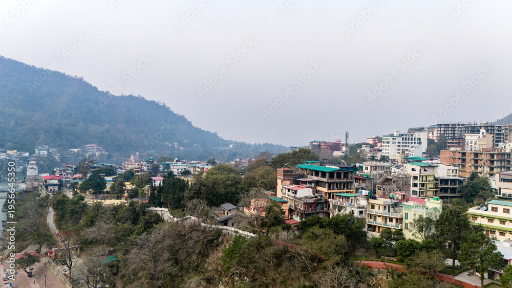 Obraz premium Aerial view of the calm Ganga River in Rishikesh, Uttarakhand on a winter morning, with misty mountains in the background and temples along the riverbank. 